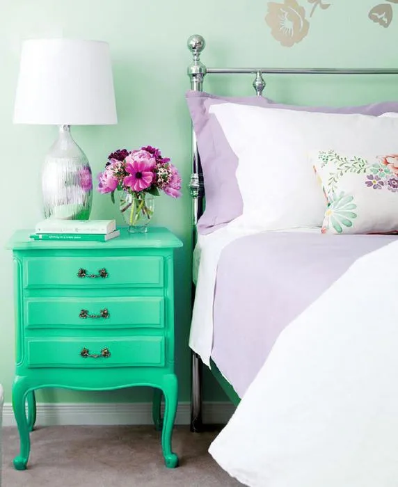 Сиреневый текстиль и прикроватная тумбочка мятного окраса в спальне частного дома